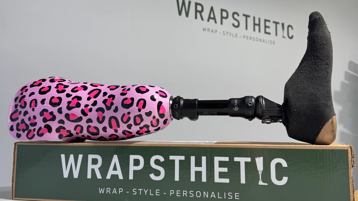 Pink leopard print Wrapsthetic prosthetic leg cover showcased on a branded box with the company logo in the background.webp