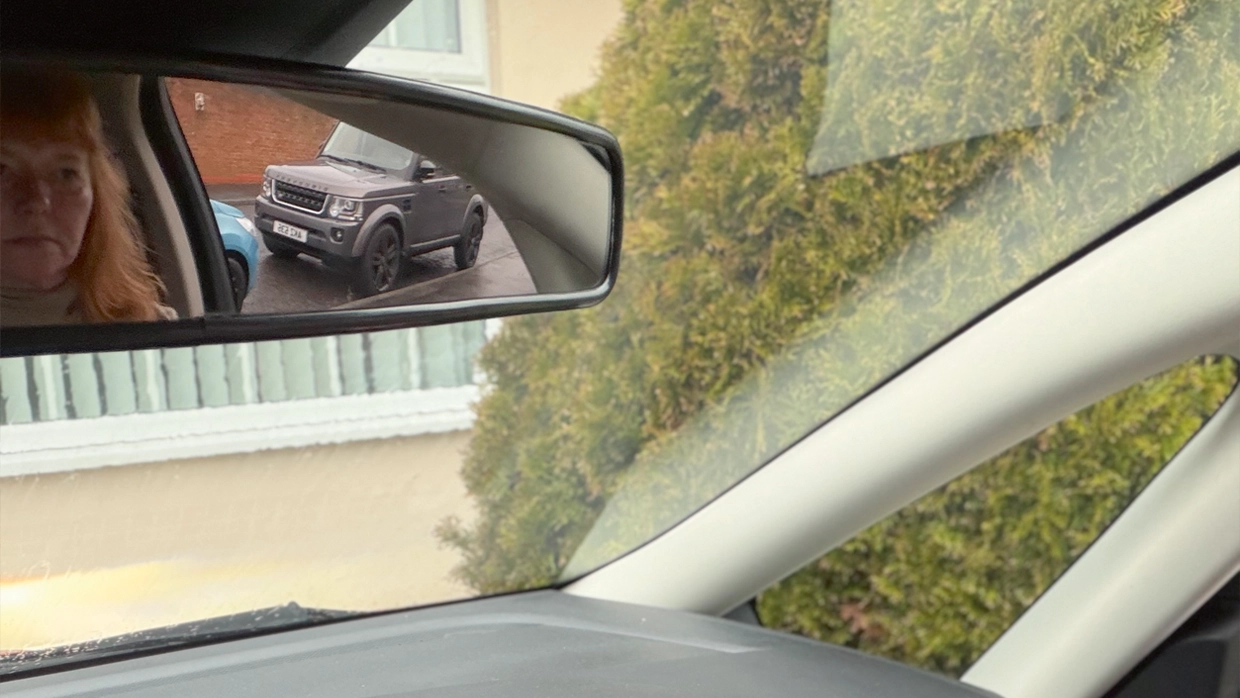 a-woman-looks-into-her-car-s-extra-wide-rearview-mirror-which-shows-a-reflection-of-her-face-and-a-grey-suv-parked-behind.webp