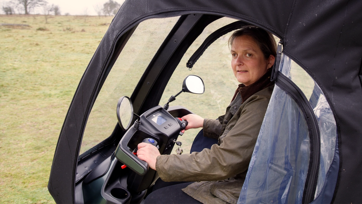 a-woman-operating-an-all-terrain-mobility-scooter-with-a-weather-proof-cab-in-a-grassy-outdoor-field.webp