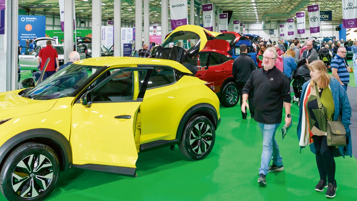 Visitors browsing various cars, including a bright yellow hatchback, at a Motability Scheme event in a large exhibition hall