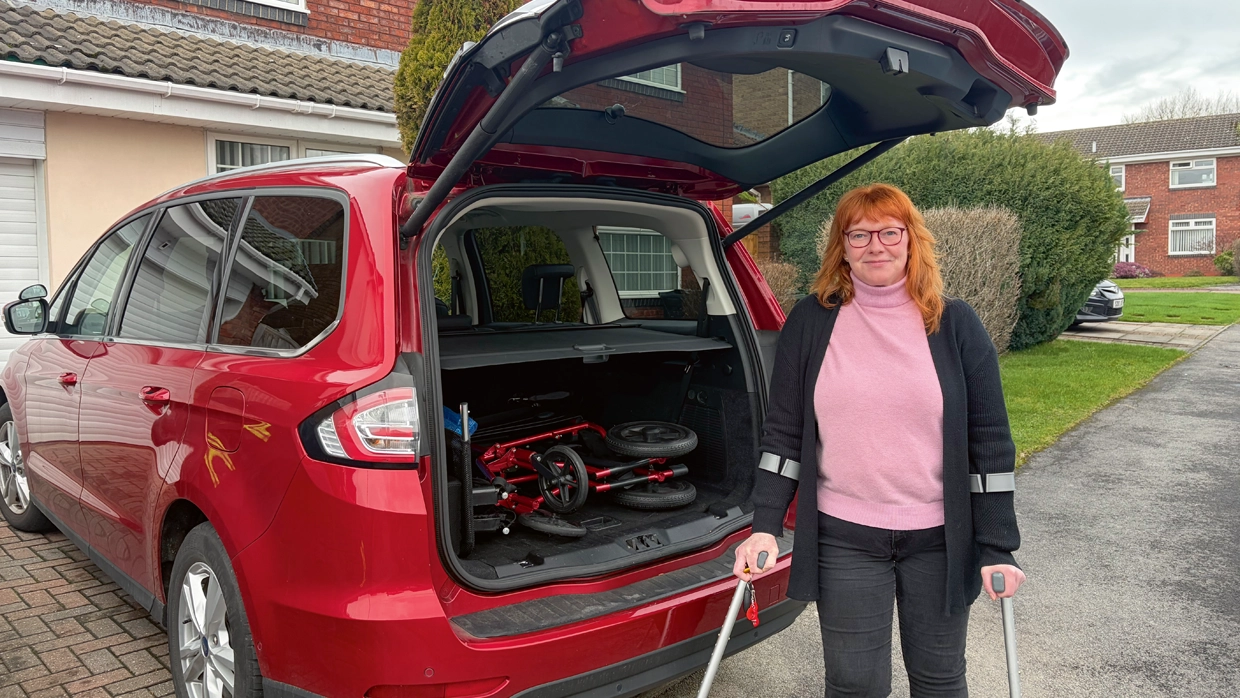 A woman using crutches stands by her red car with an open boot containing a folded mobility walker