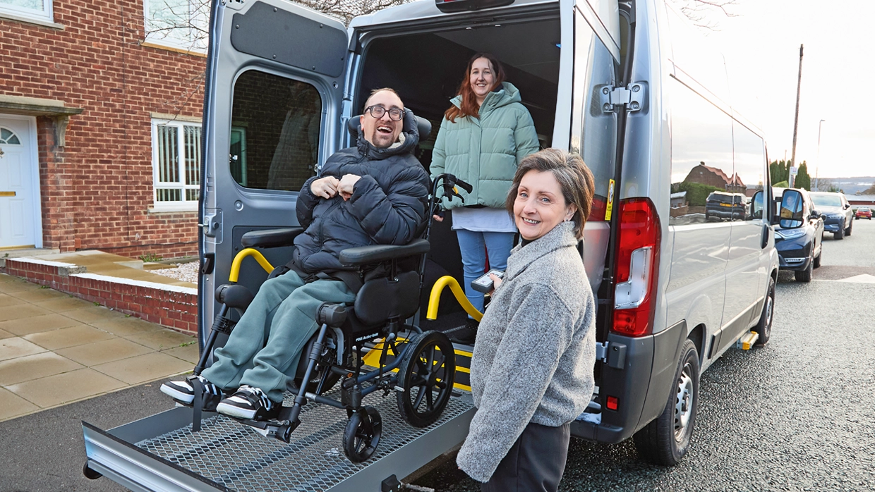 A male wheelchair user in his 30s posing outside on the tailgate lift of a large WAV, with a young woman standing behind him and his mother standing in the foreground.