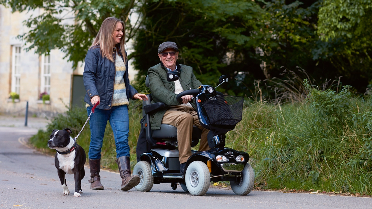 A smiling man on a mobility scooter enjoys an outdoor walk with a companion and their dog along a tree-lined path.