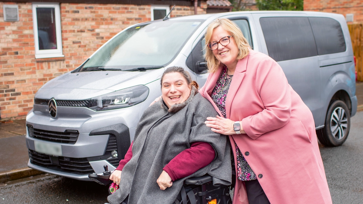 A smiling woman in a power wheelchair and her mother pose in front of a silver Citroën wheelchair accessible van.