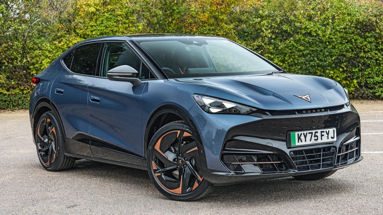 A metallic blue Cupra Tavascan parked on asphalt in front of tall hedgerow