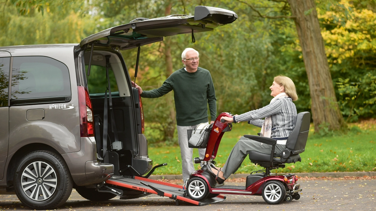 A female pensioner on a mobility scooter uses a rear ramp to enter a grey Wheelchair Accessible Vehicle (WAV) while an elderly man assists.
