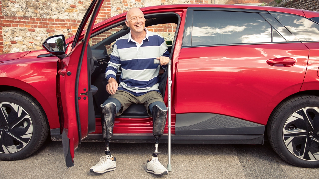 A smiling man with bilateral prosthetic legs sits in the driver's seat of a red car with a walking stick, highlighting mobility independence.