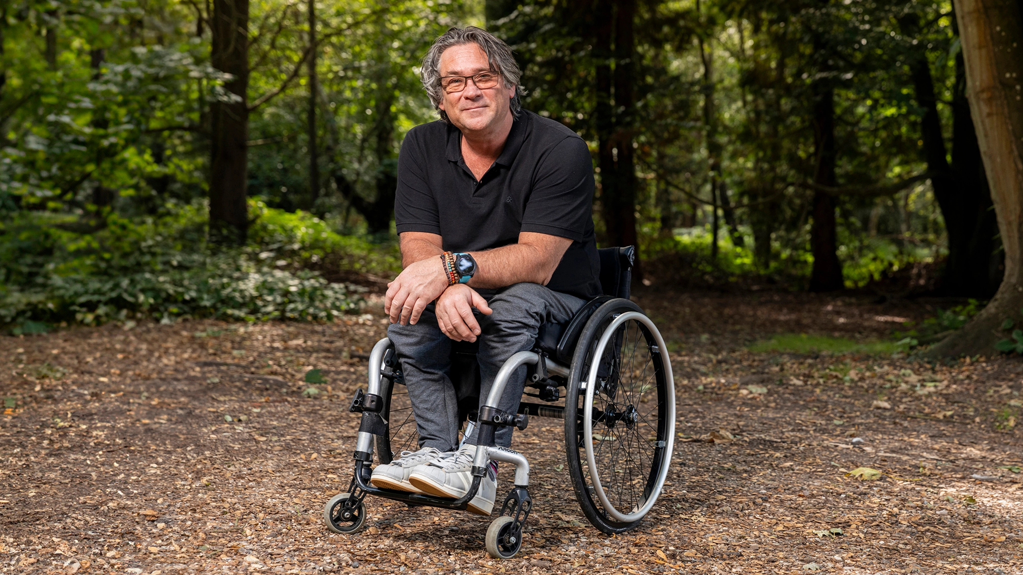 Grant sitting in his wheelchair in a woodland clearing, wearing a black polo and dark jeans smiling