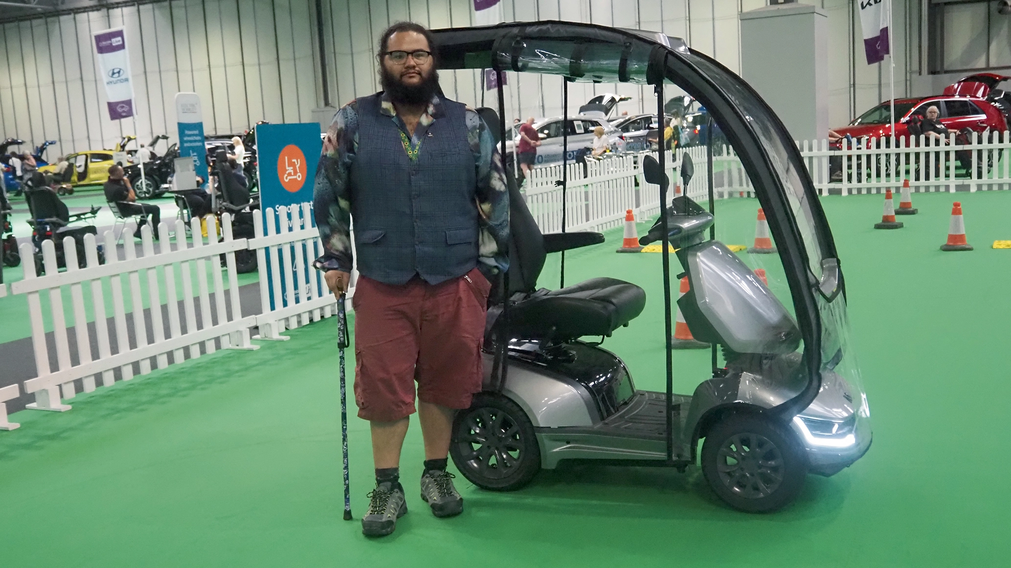 A man, stood holding a walking stick, next to a canopied mobility scooter