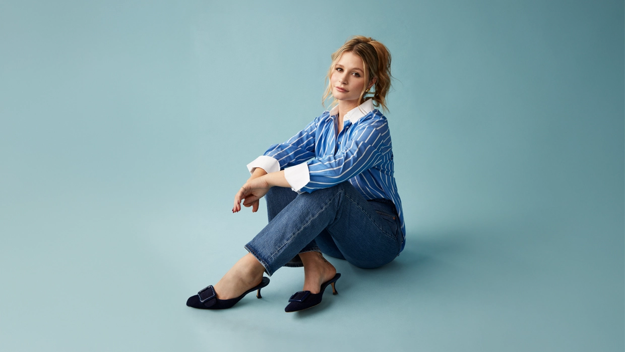 Photo of Mollie Pearce sat down in front of a blue background. She is wearing jeans, a shirt and heels. 