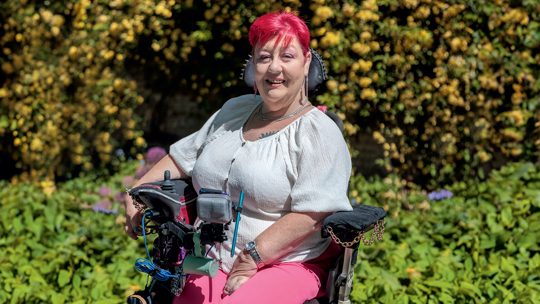 Caroline has pink hair, and she is sat on her wheelchair smiling at the camera