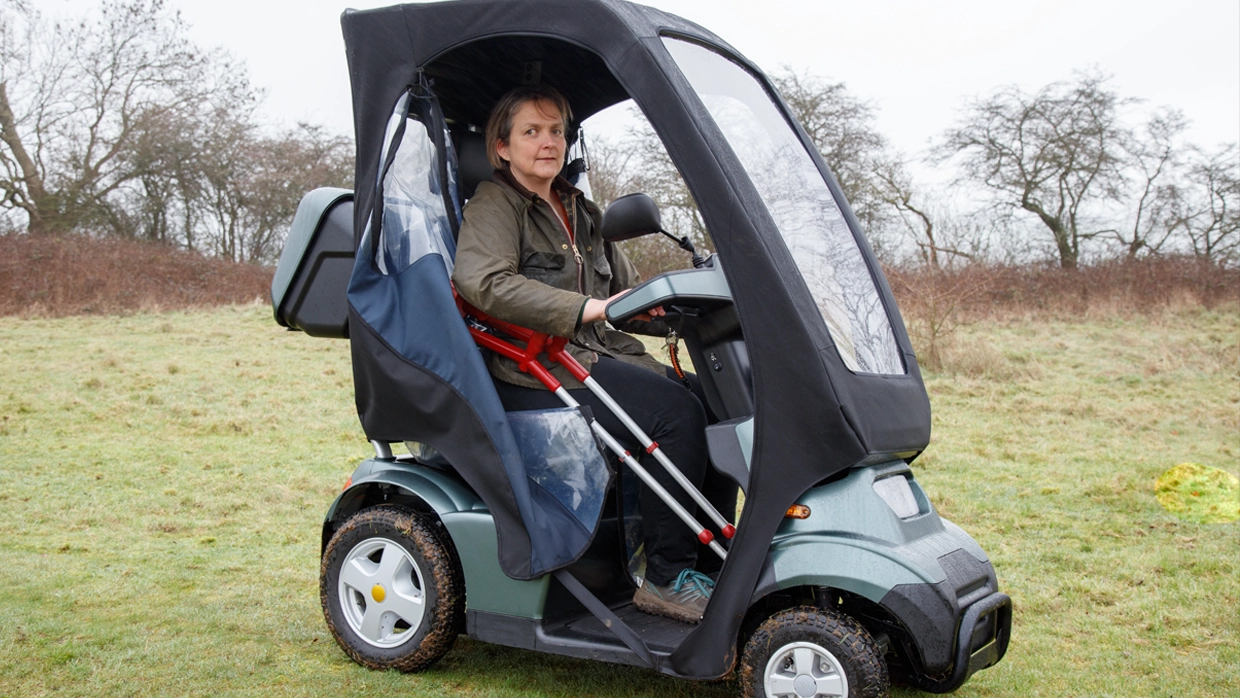 A woman in an all-terrain mobility scooter with a weather-proof cab and crutch holder, outdoors in a grassy field