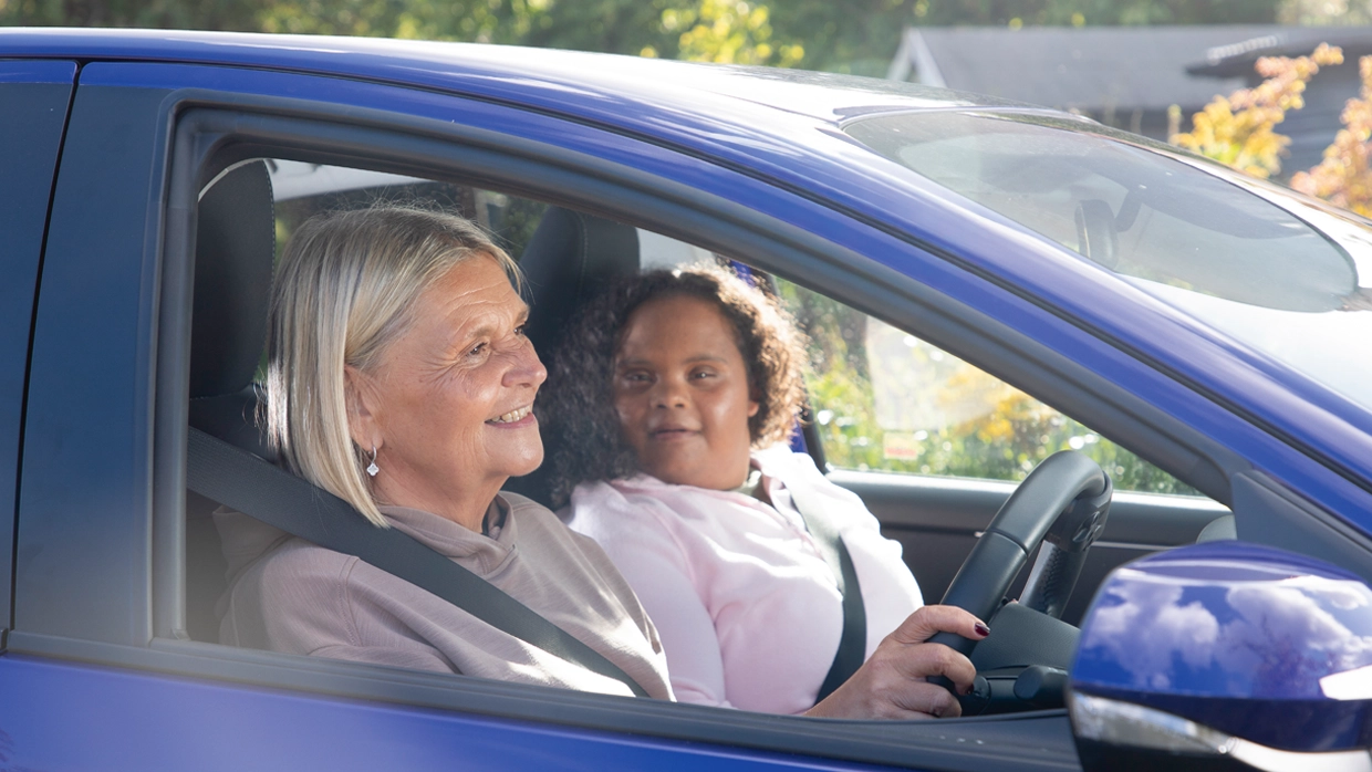 A woman smiles from the driver's seat of a blue car next to an older passenger.