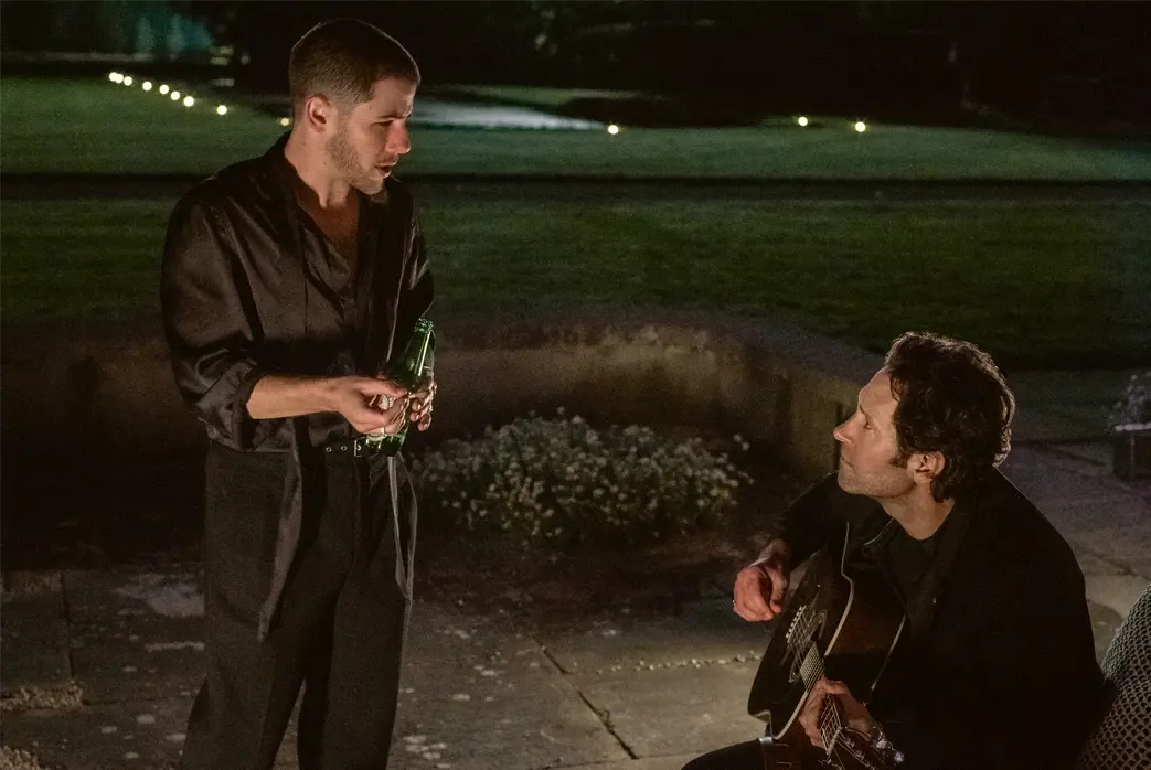 Nick Jonas stands holding a bottle while Paul Rudd plays an acoustic guitar in a nighttime scene from the film 'Power Ballad'