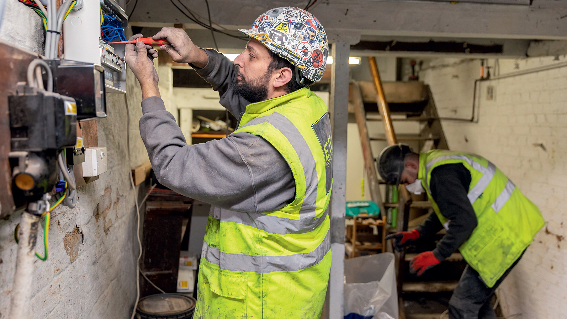 one-deaf-builder-wiring-electrics-and-another-in-background-of-basement-looking-in-a-bin.jpg