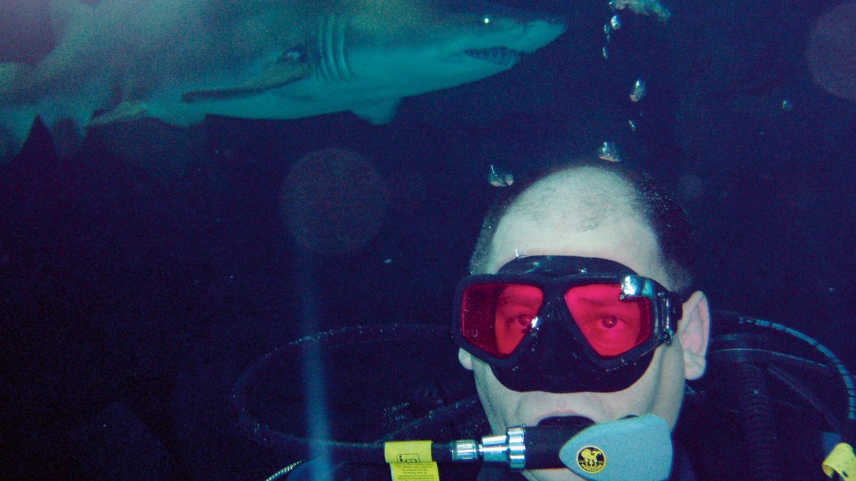 Fraser scuba diving with a shark behind him.webp