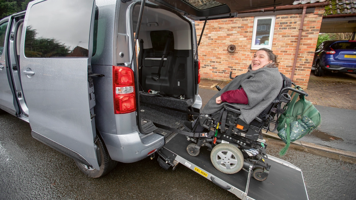a-smiling-woman-in-a-power-wheelchair-uses-a-rear-entry-ramp-to-board-a-silver-wheelchair-accessible-vehicle-wav.webp